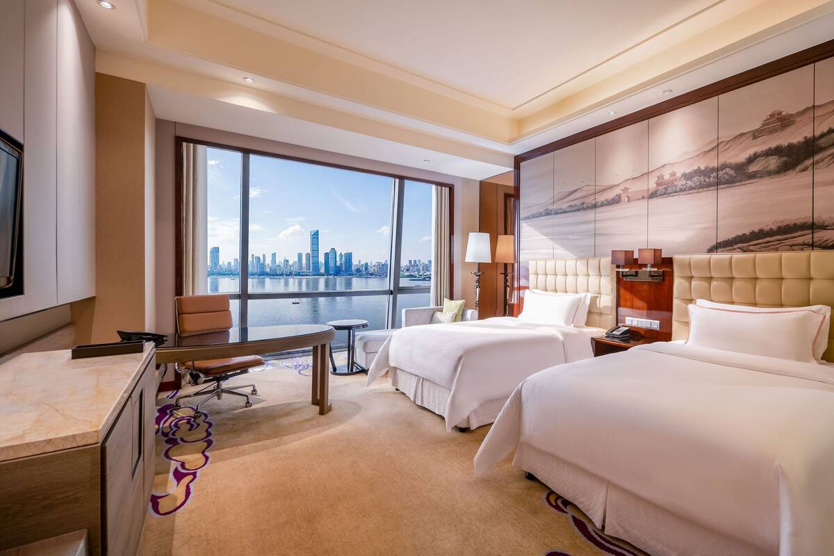 Habitación de hotel con dos camas, un escritorio, una silla y un amplio ventanal con vista a la ciudad.