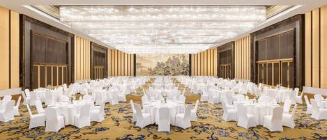 Un salón de banquetes con mesas redondas dispuestas para un evento formal, con una alfombra estampada y una lámpara de candelabros grande.