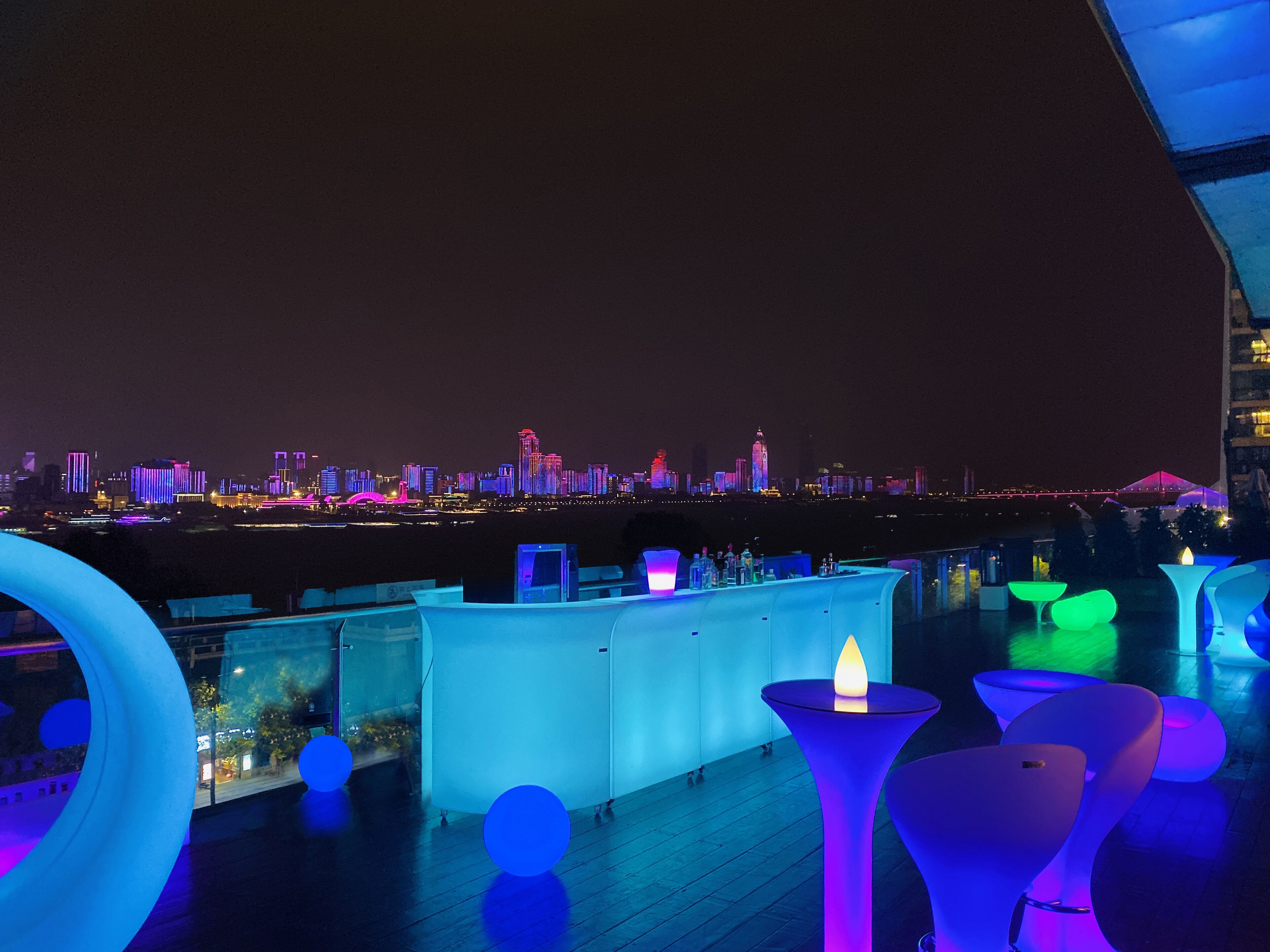 Um bar no terraço com assentos modernos e vista para o horizonte urbano à noite.