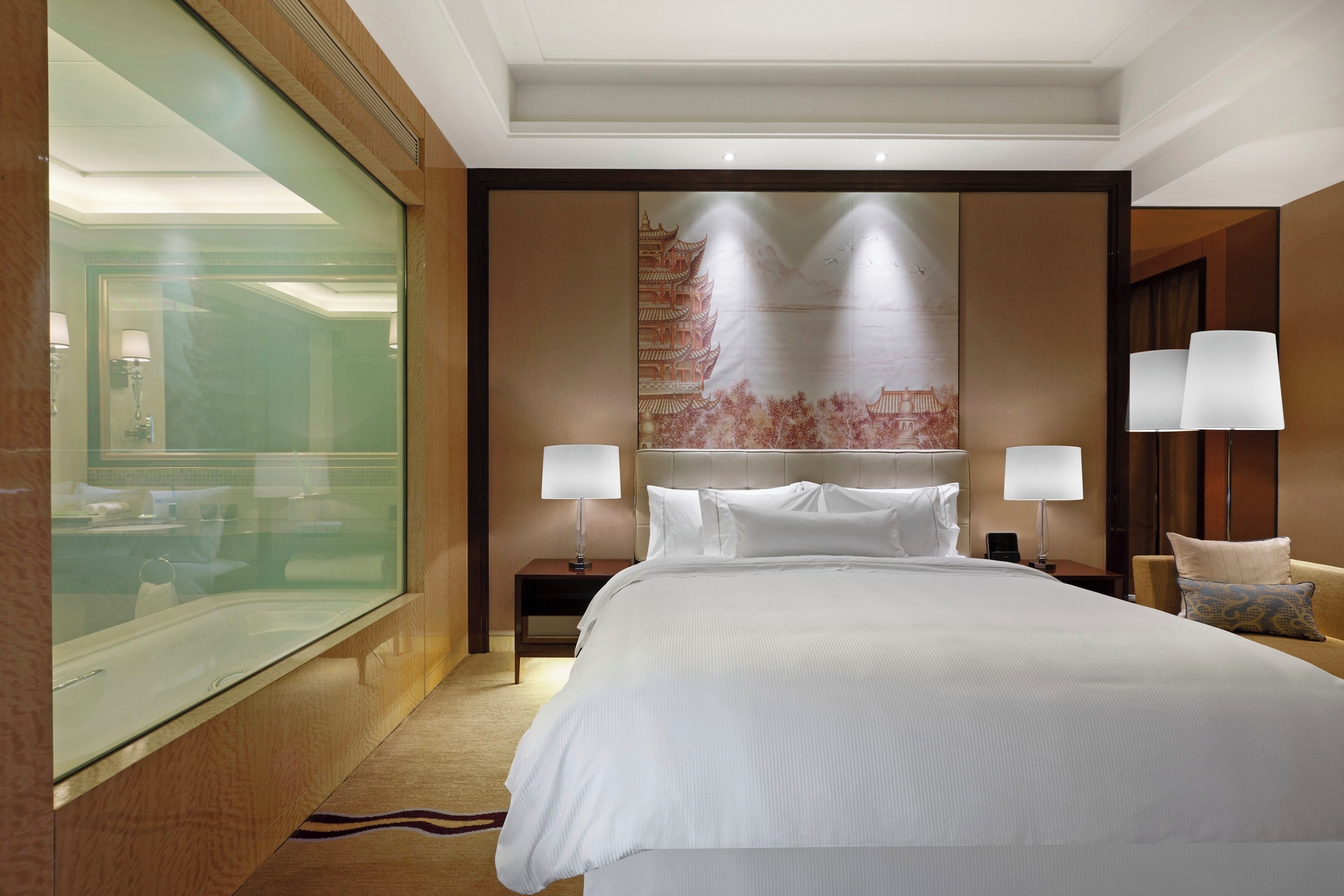 Habitación de hotel con una cama grande, un separador de vidrio, un mural decorativo, dos lámparas de noche y una mesita de noche pequeña.
