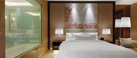 Habitación de hotel con una cama grande, un separador de vidrio, un mural decorativo, dos lámparas de noche y una mesita de noche pequeña.