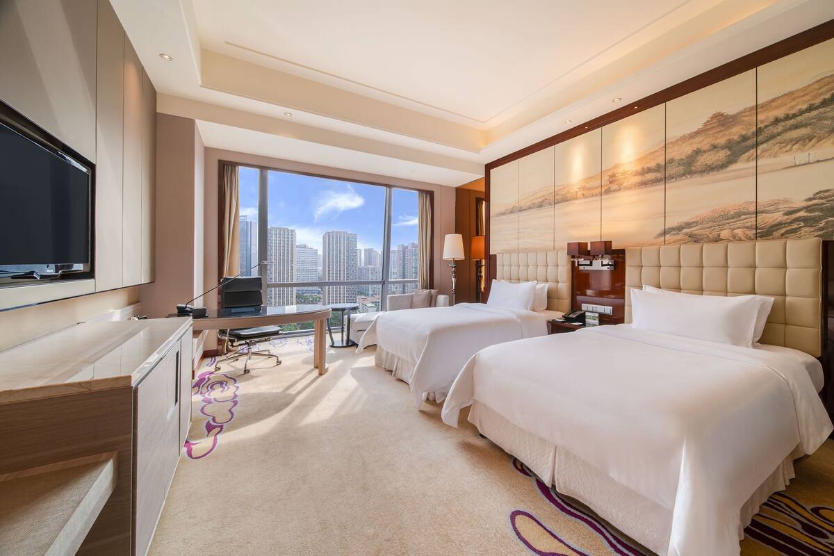 Habitación de hotel con dos camas, un escritorio, un televisor y un ventanal con vistas a la ciudad.