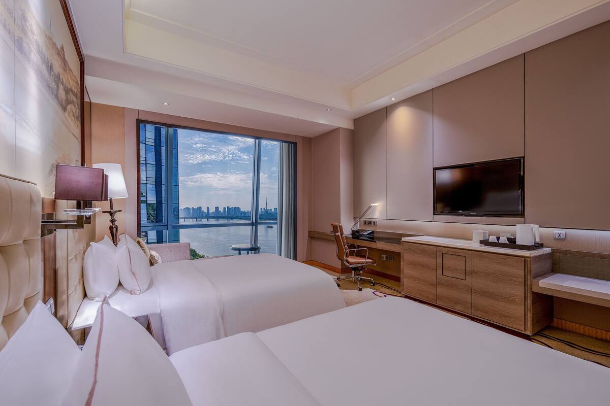 Habitación de hotel con dos camas, un escritorio, un televisor y vistas a un horizonte urbano.