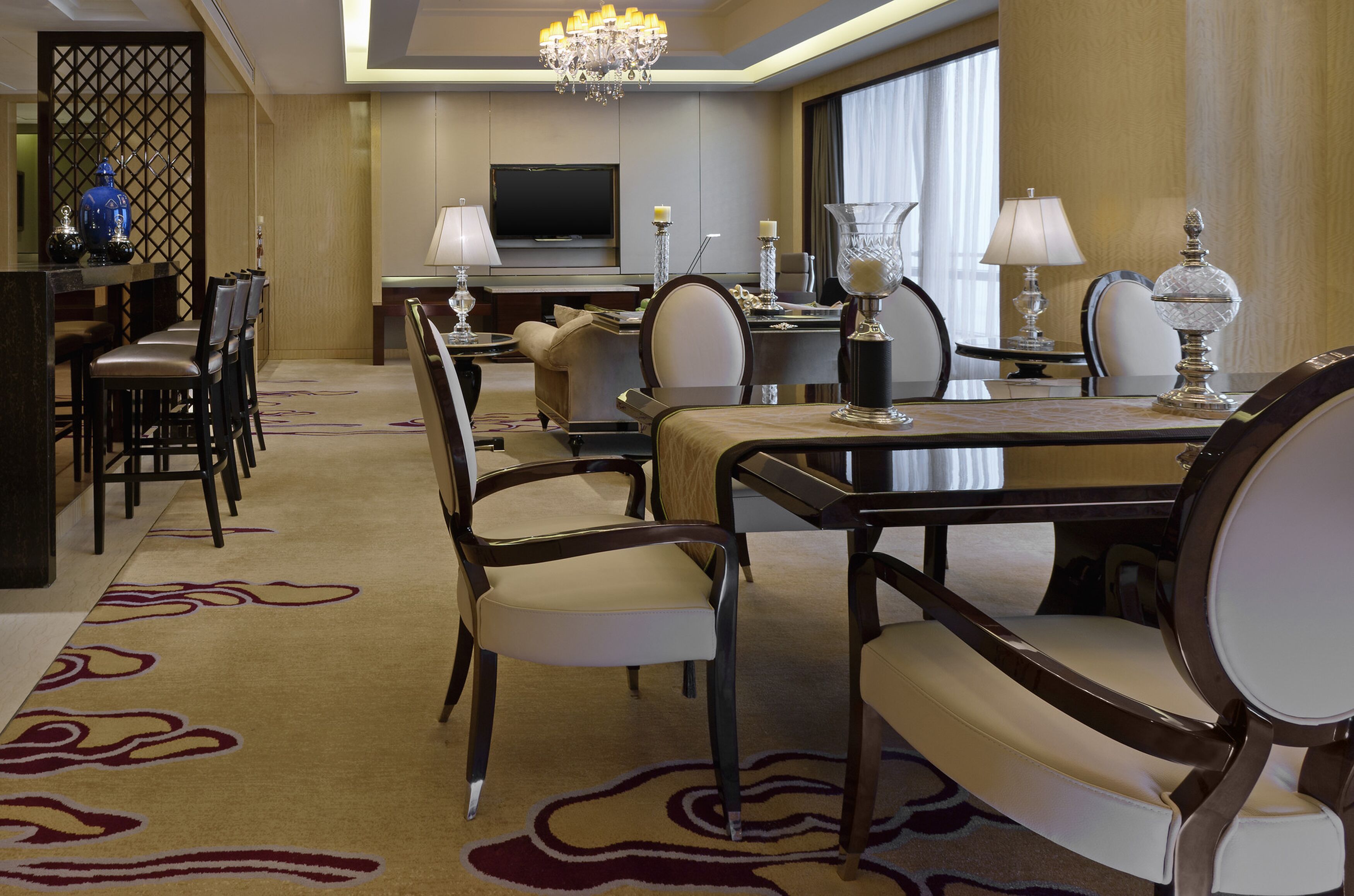 Uma sala de jantar de hotel com uma mesa comprida, cadeiras, um candelabro e uma televisão ao fundo.