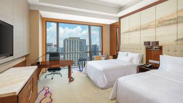 A hotel room with a large window, two beds, a desk, and a TV.