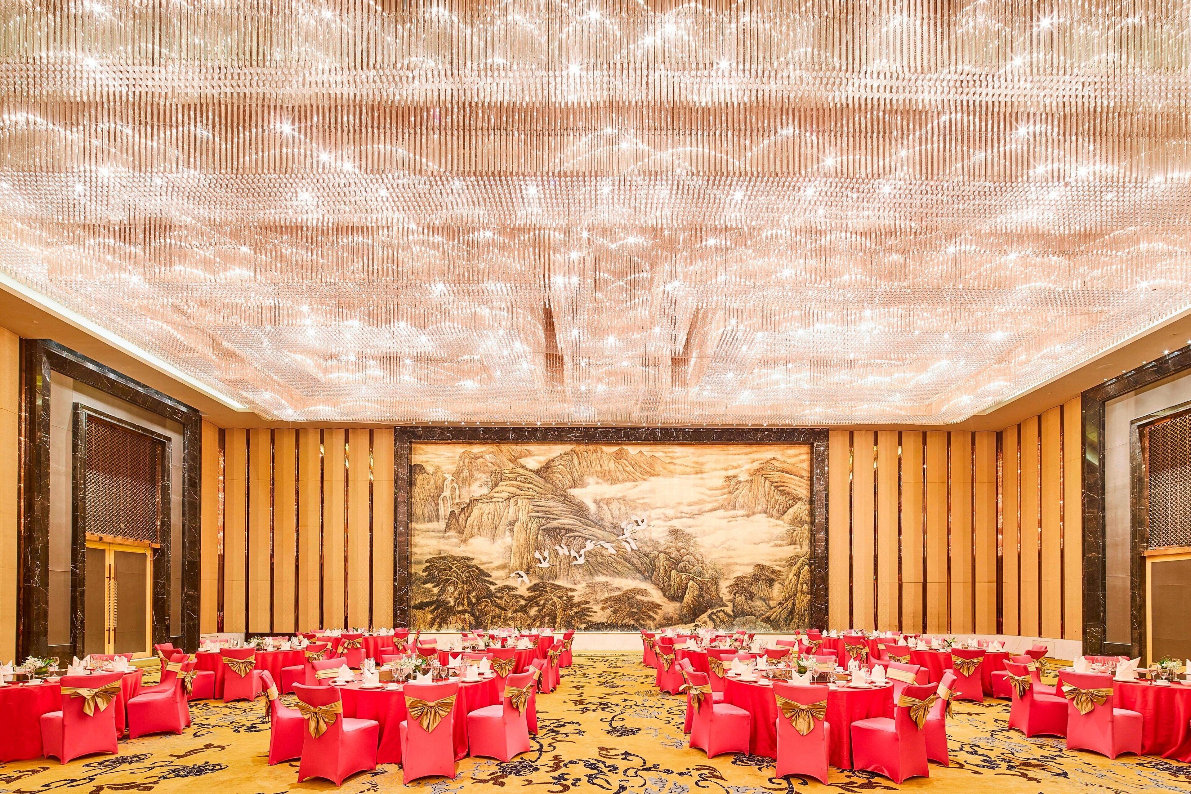 A banquet hall with rows of tables set for a formal event, featuring a large mural on the back wall and a ceiling with a light fixture.