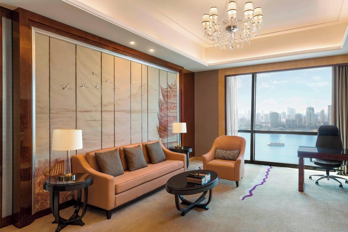 Habitación de hotel moderna con sofá, sillones, escritorio y un amplio ventanal con vista a la ciudad.