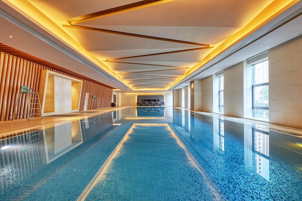 Una piscina interior larga y estrecha con suelo reflectante, paneles de madera en un lateral y amplias ventanas al otro lado.