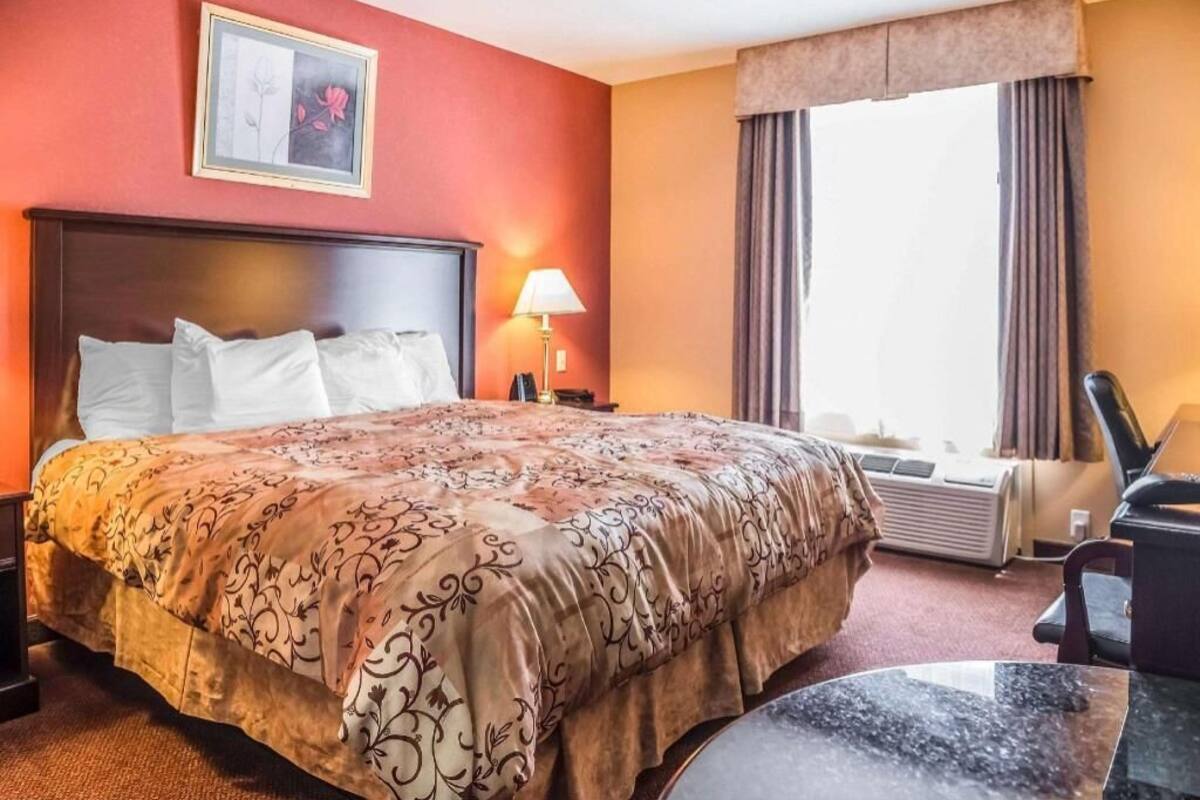 Una habitación de hotel con una cama grande, una mesita de noche con lámpara, una silla y una ventana con cortinas.