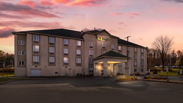 Ein mehrgeschossiges Hotel mit zentralem Eingang, einem Parkplatz und wolkenüberschwemmtem Himmel.