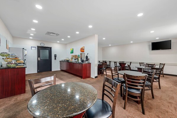 A hotel room with a kitchen area, dining table, chairs, and a TV mounted on the wall.