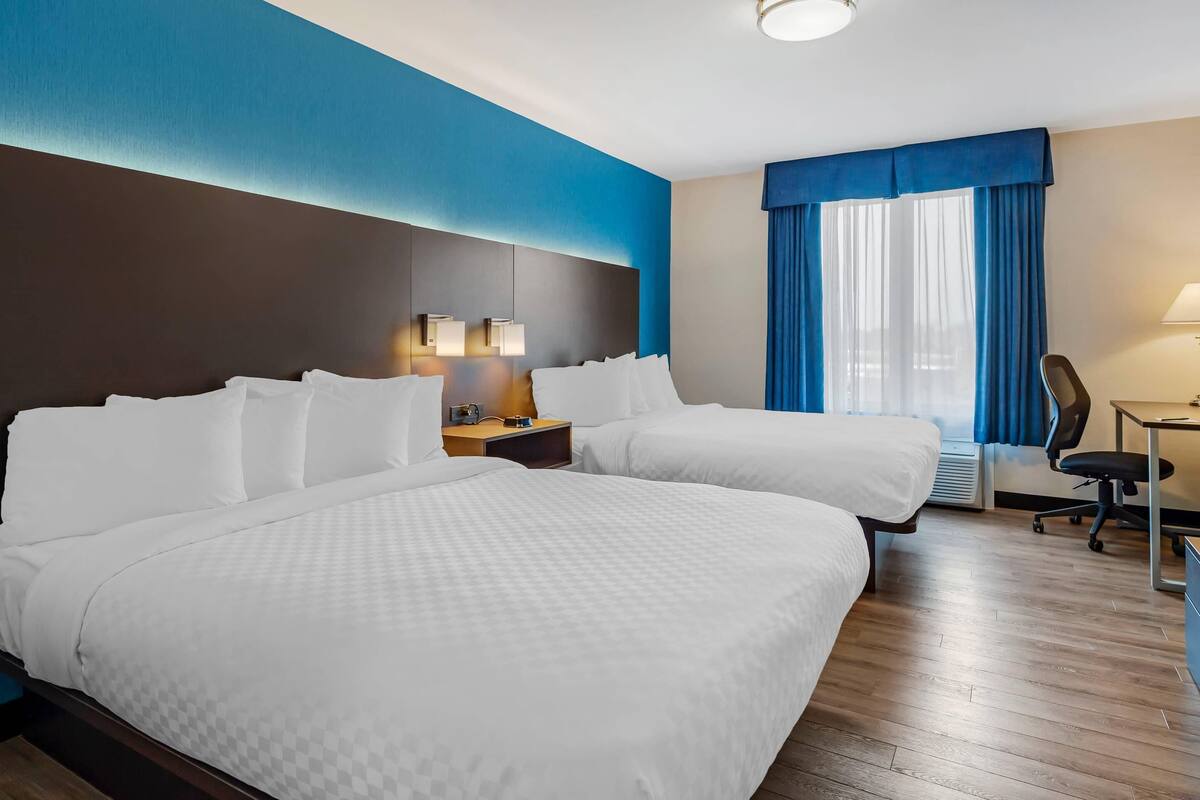 Habitación de hotel con dos camas, un escritorio, una silla, una mesita de noche y una ventana con cortinas azules.