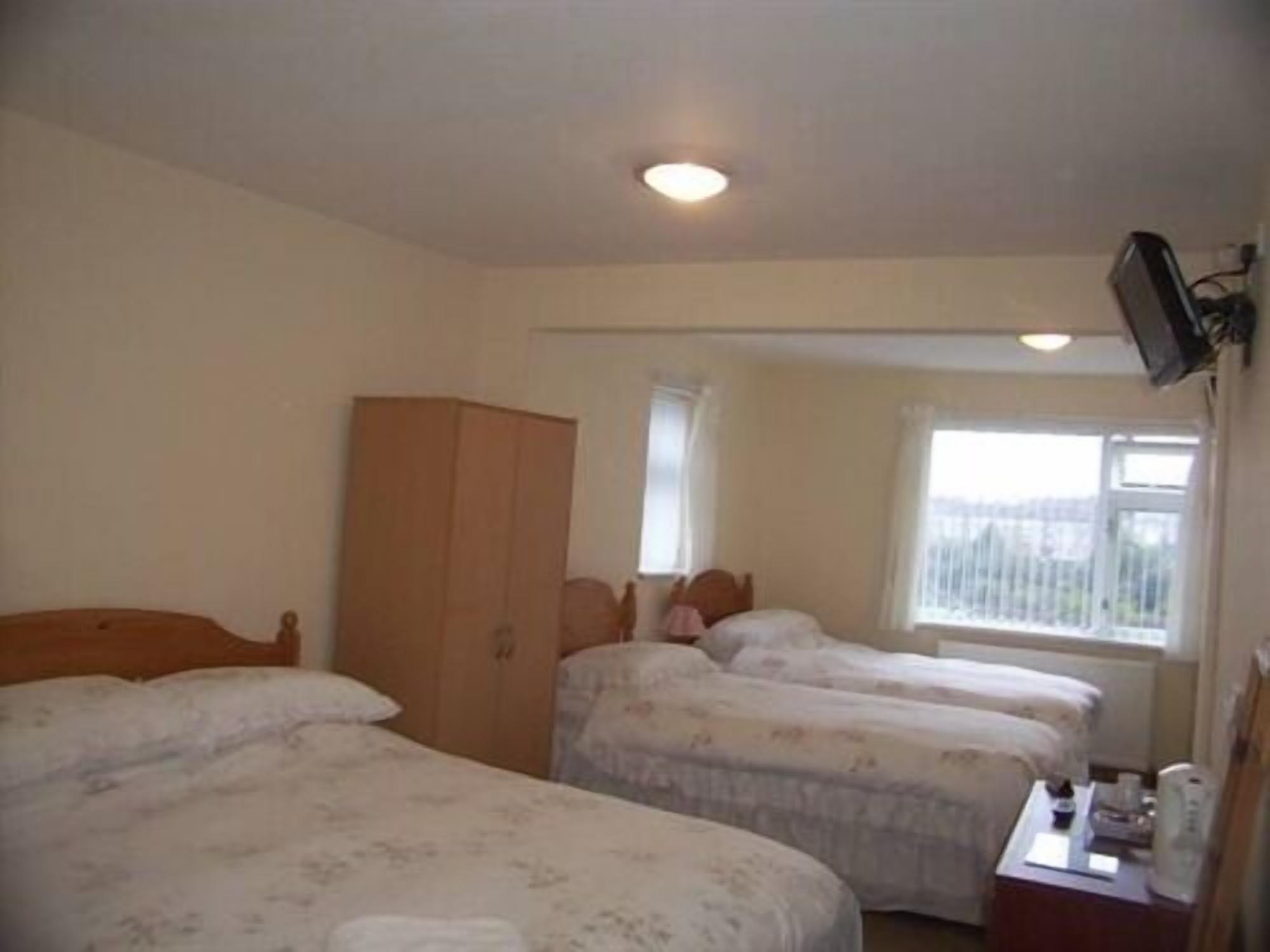 A hotel room with three single beds, a wooden wardrobe, a television mounted on the wall, and a window providing a view of the outside.