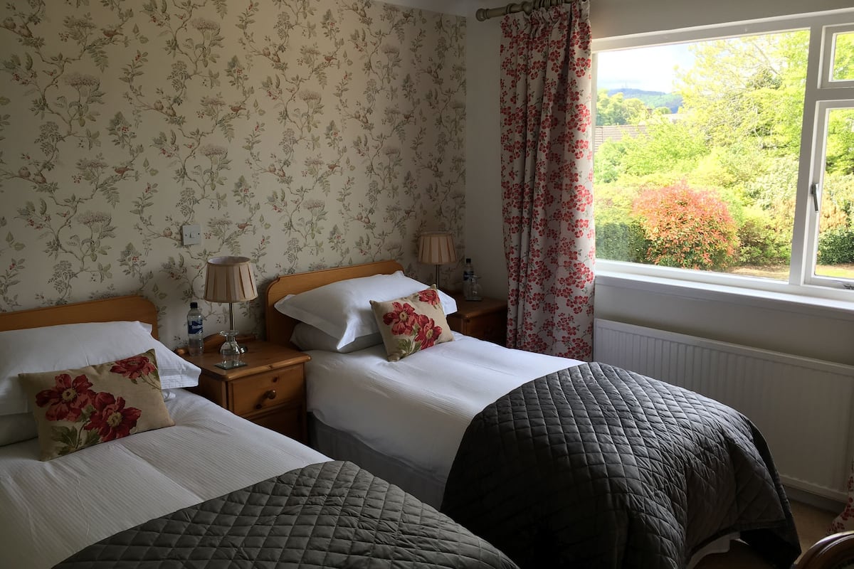 A room with two beds, floral wallpaper, a window with a view of greenery, and a radiator.