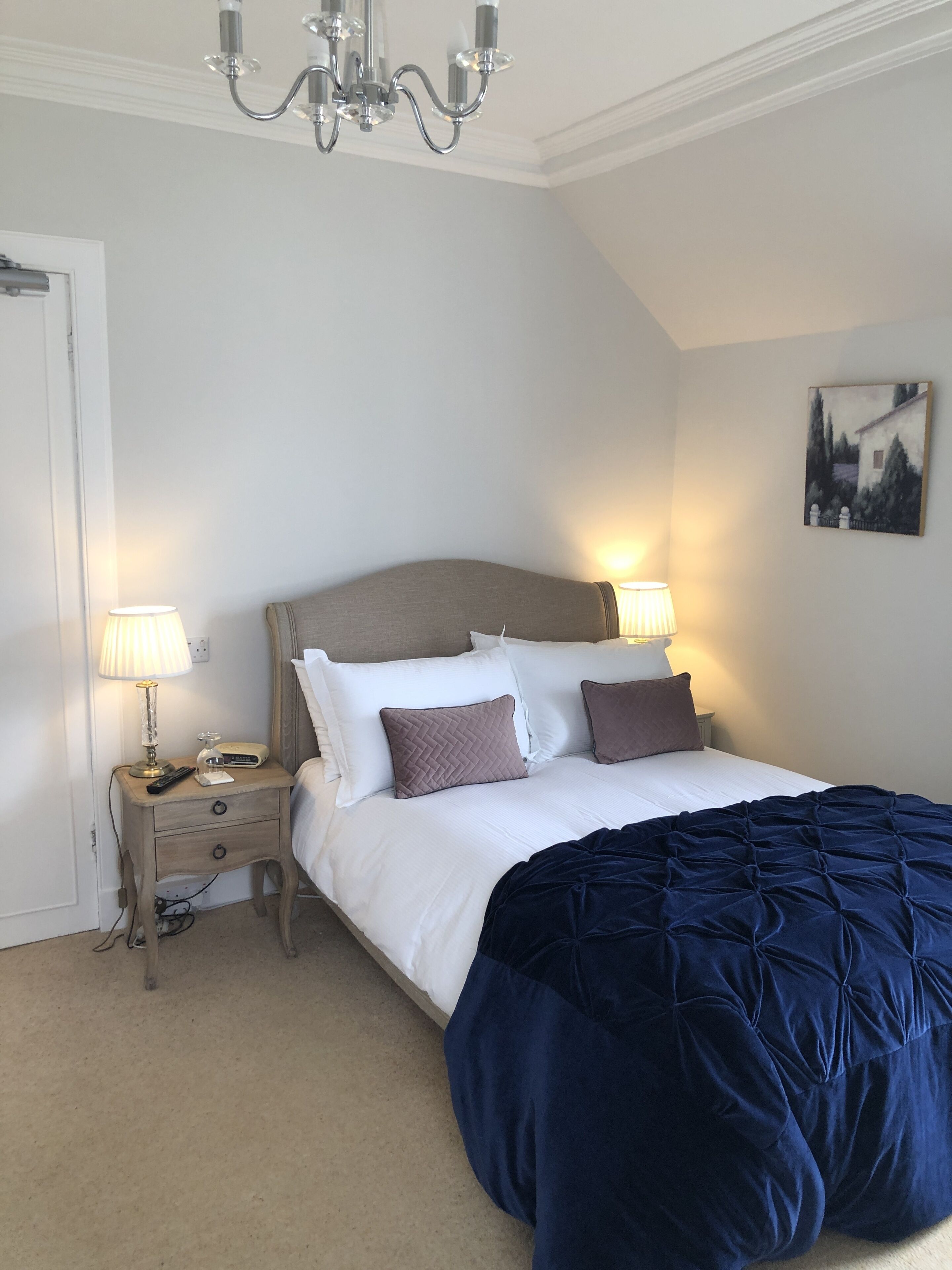 A bedroom with a bed, two bedside lamps, a nightstand, a chandelier, and a painting on the wall.