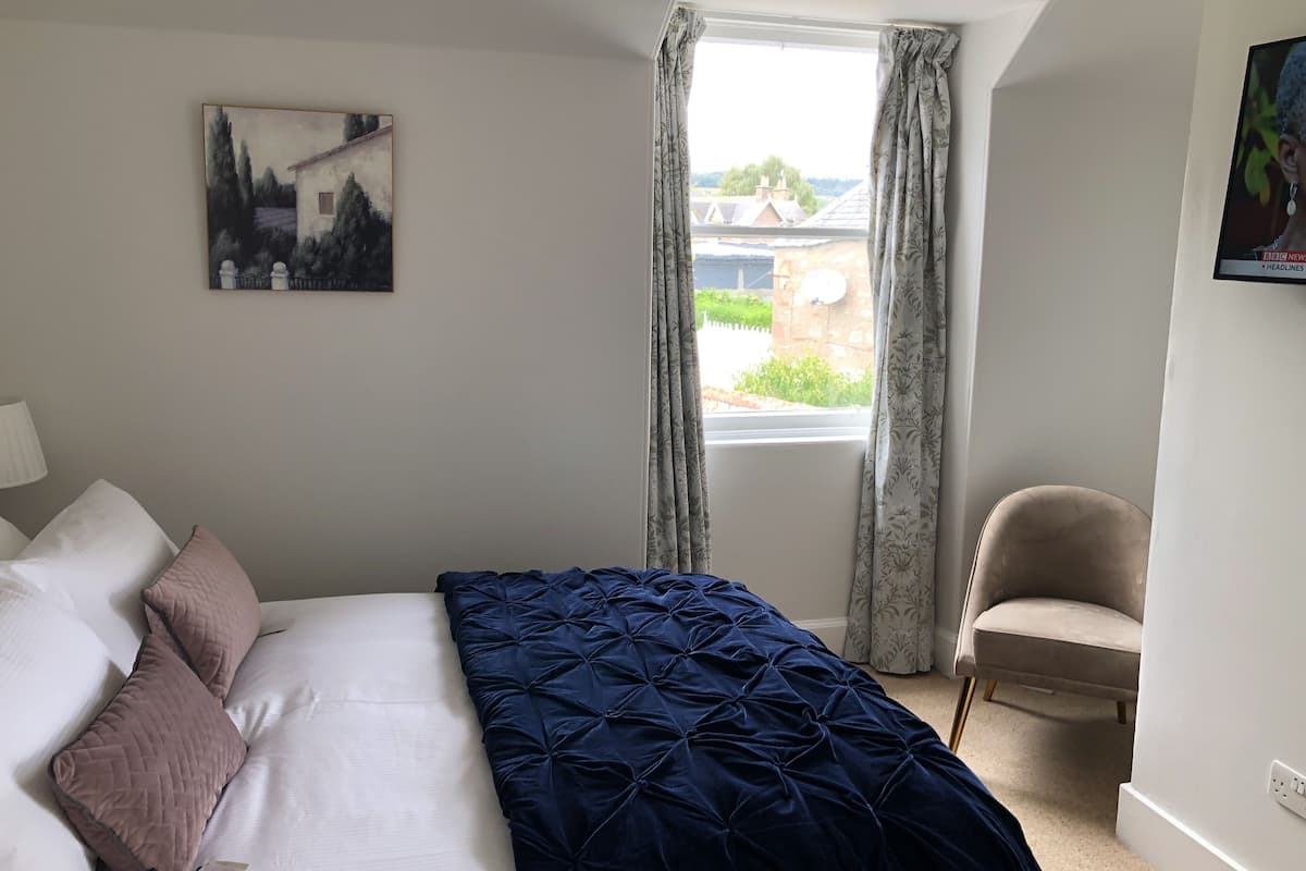 A bedroom with a bed, a chair, a window with curtains, and a painting on the wall.