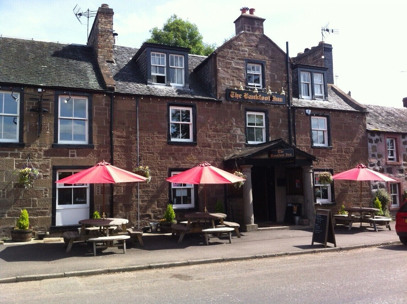 Un bâtiment traditionnel en pierre, sur lequel on peut lire l’inscription « The Bankfoot Inn ». Il y a des tables à l’extérieur et des parasols rouges.