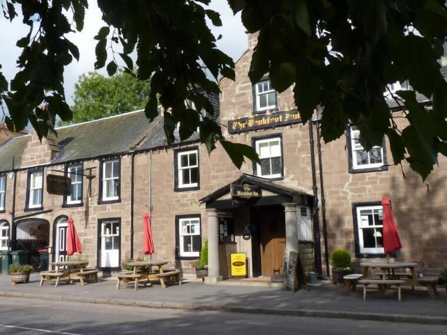 Un bâtiment traditionnel en pierre, avec une enseigne de pub, des tables à l’extérieur et un panneau indicateur.