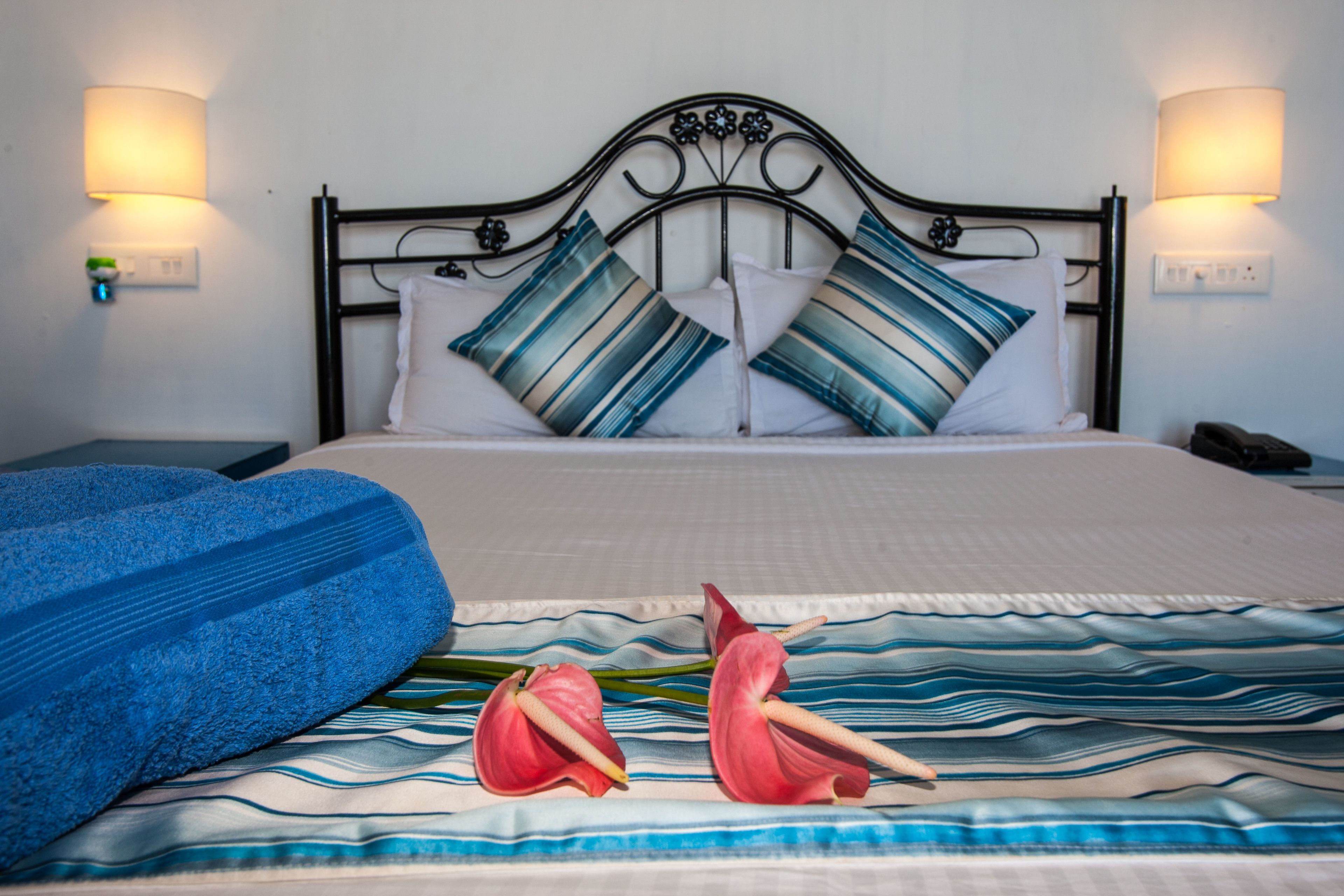 Un lit bien fait, avec des oreillers à rayures, une couverture bleue et deux fleurs rouges posées sur le couvre-lit.