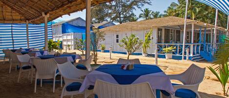 An outdoor seating area with white wicker chairs and blue tablecloths, a covered patio, and a building with a blue and white color scheme.
