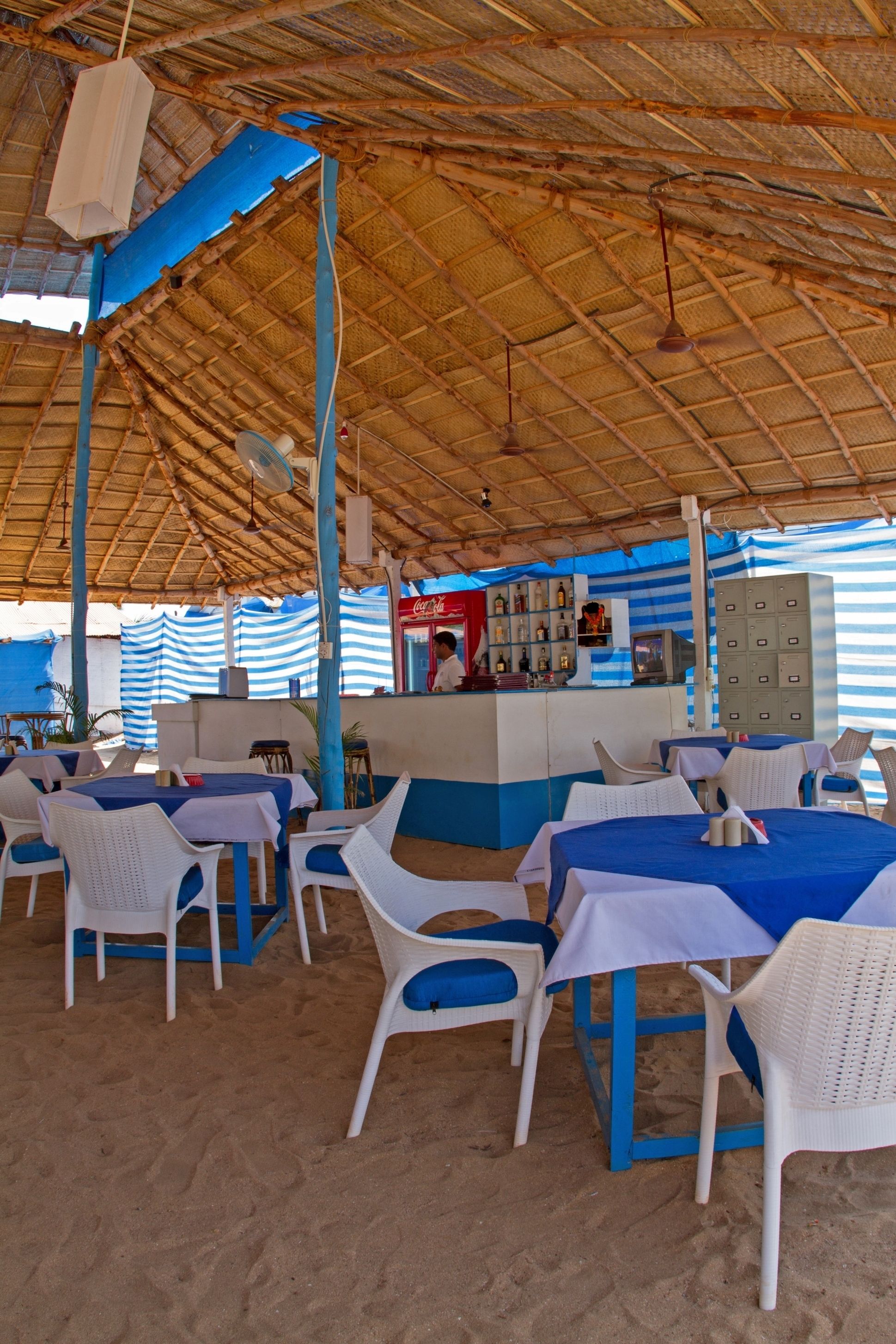 Un restaurant en plein air avec des chaises et des tables en plastique blanc, un auvent à rayures bleues et un réfrigérateur Coca-Cola.