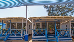 A row of beach huts with blue and white striped awnings, each with a small seating area and a thatched roof. - Cuba Agonda (Canacona)