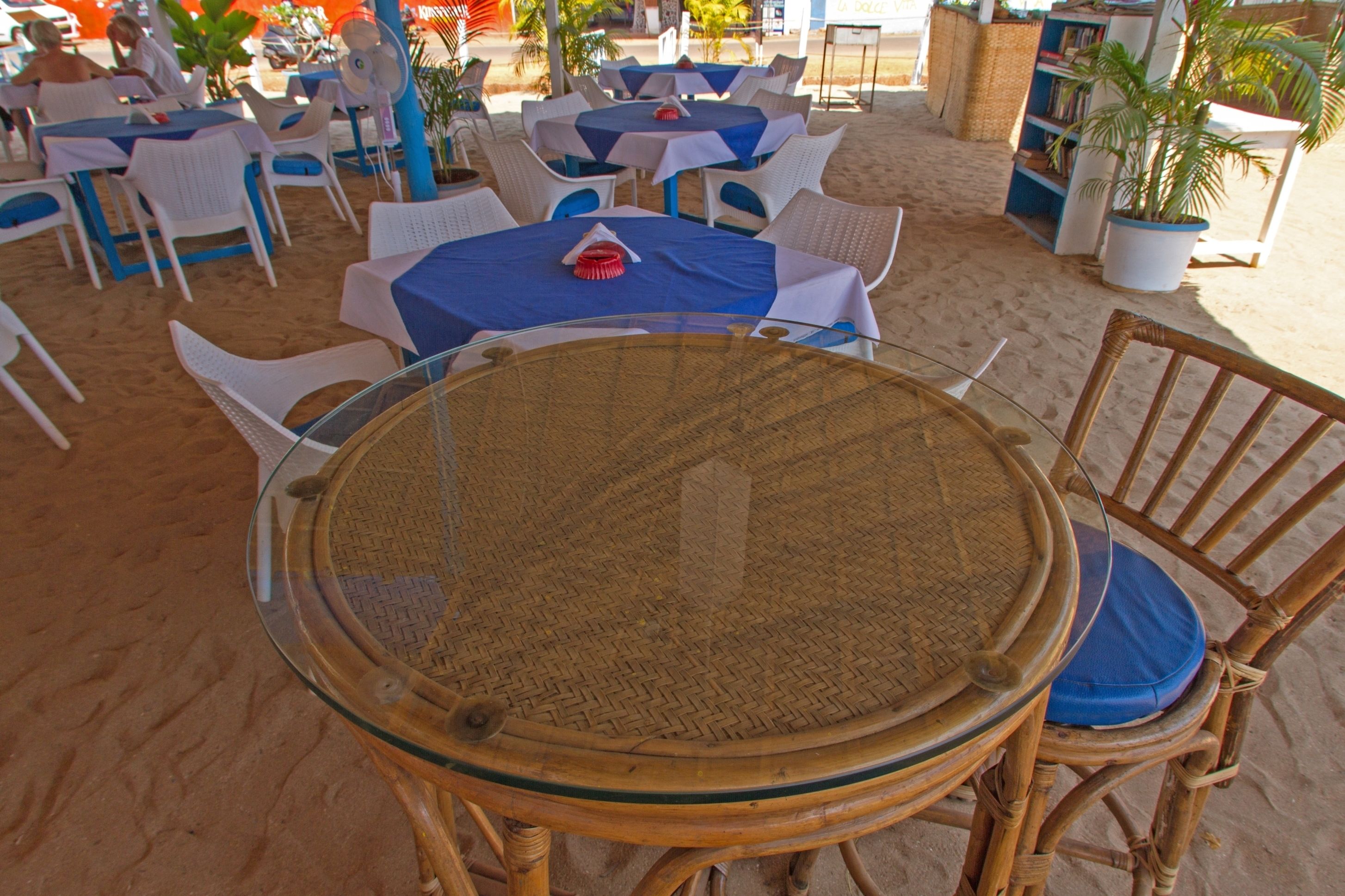 A beachside restaurant with round tables and wicker chairs, surrounded by sand.