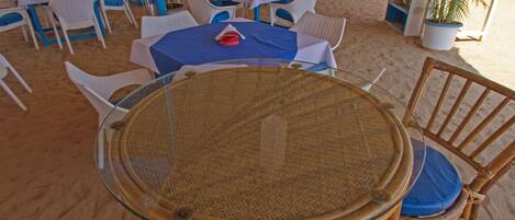 A beachside restaurant with round tables and wicker chairs, surrounded by sand.