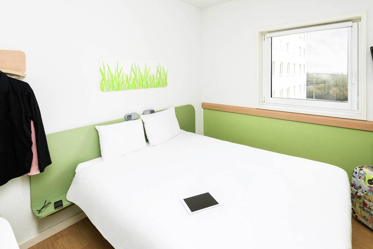 A hotel room with a bed, a window, a green headboard, a wall art of grass, and a small table with a book.