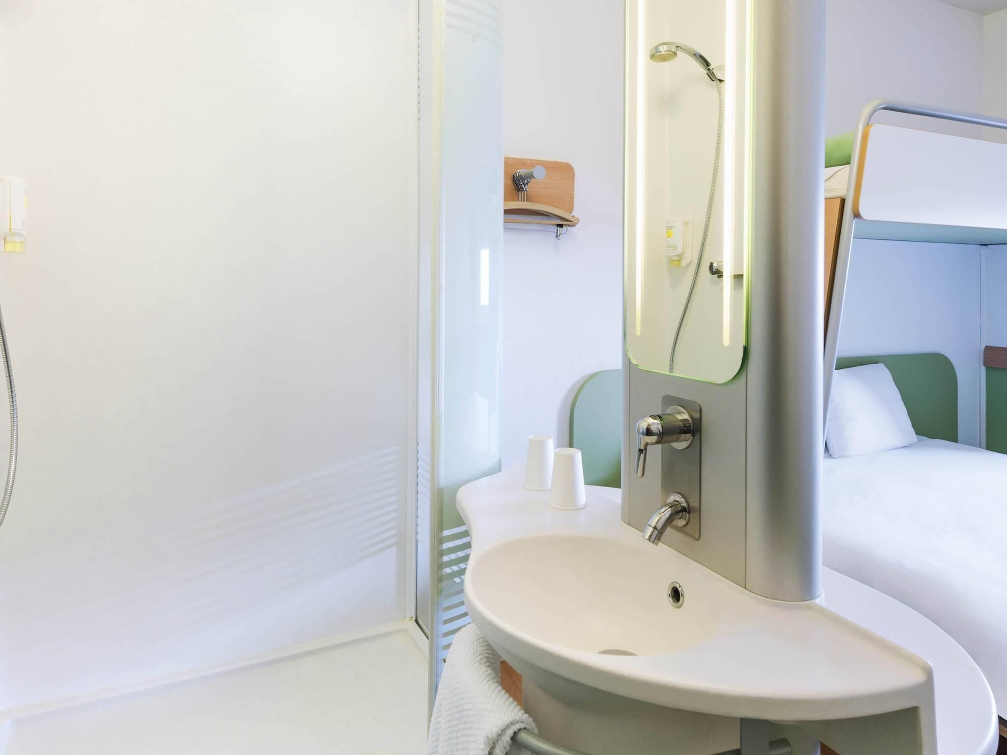 A bathroom with a sink, mirror, shower, and a bed with green headboard.
