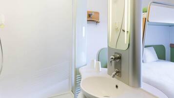 A bathroom with a sink, mirror, shower, and a bed with green headboard.