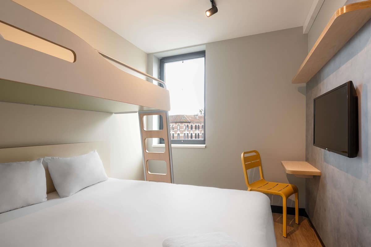 A modern hotel room with a large bed, a wooden ladder, a small desk, a chair, and a TV mounted on the wall.