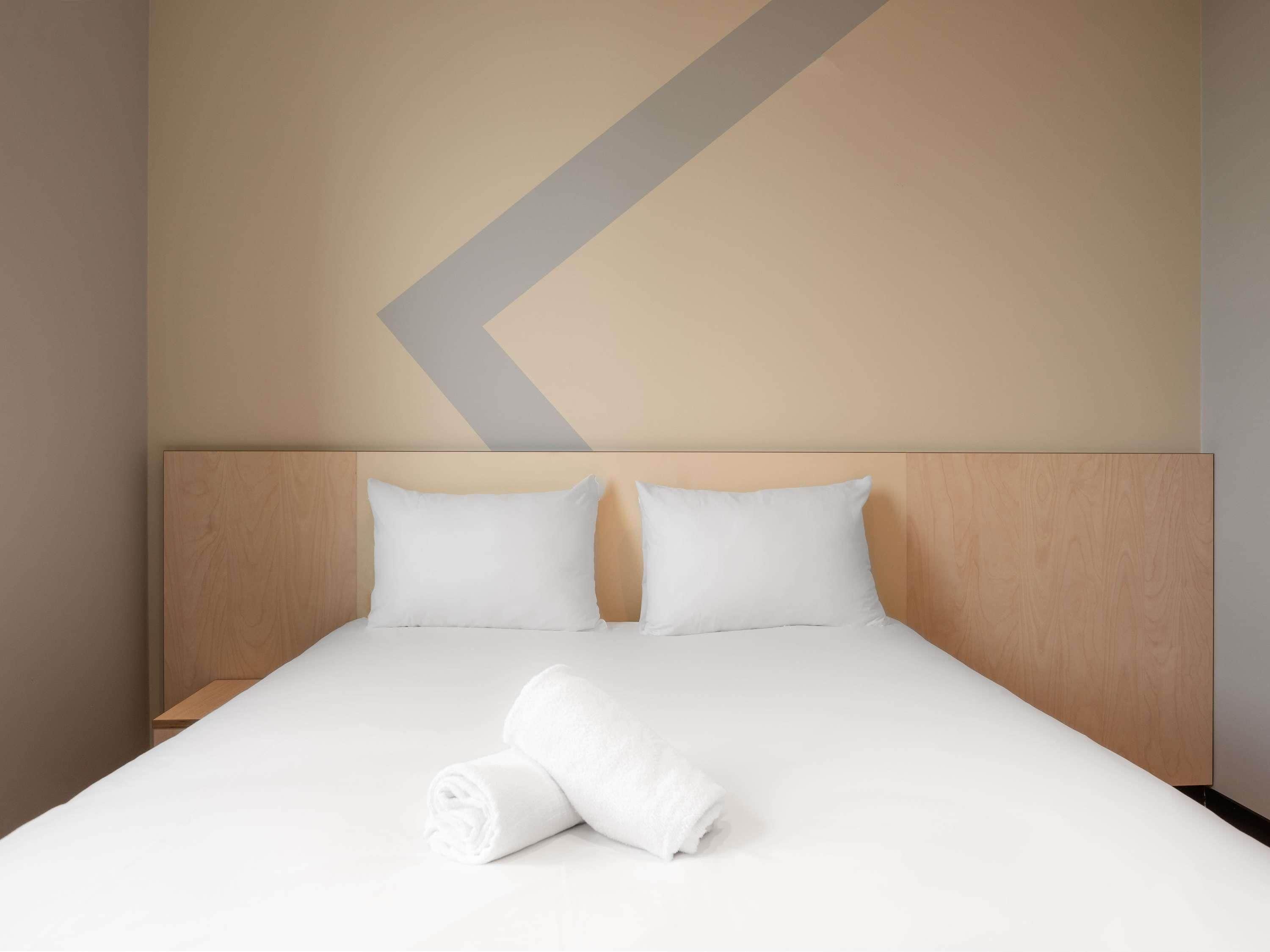 A neatly made bed with two pillows and a rolled towel, set against a wall with a geometric design.