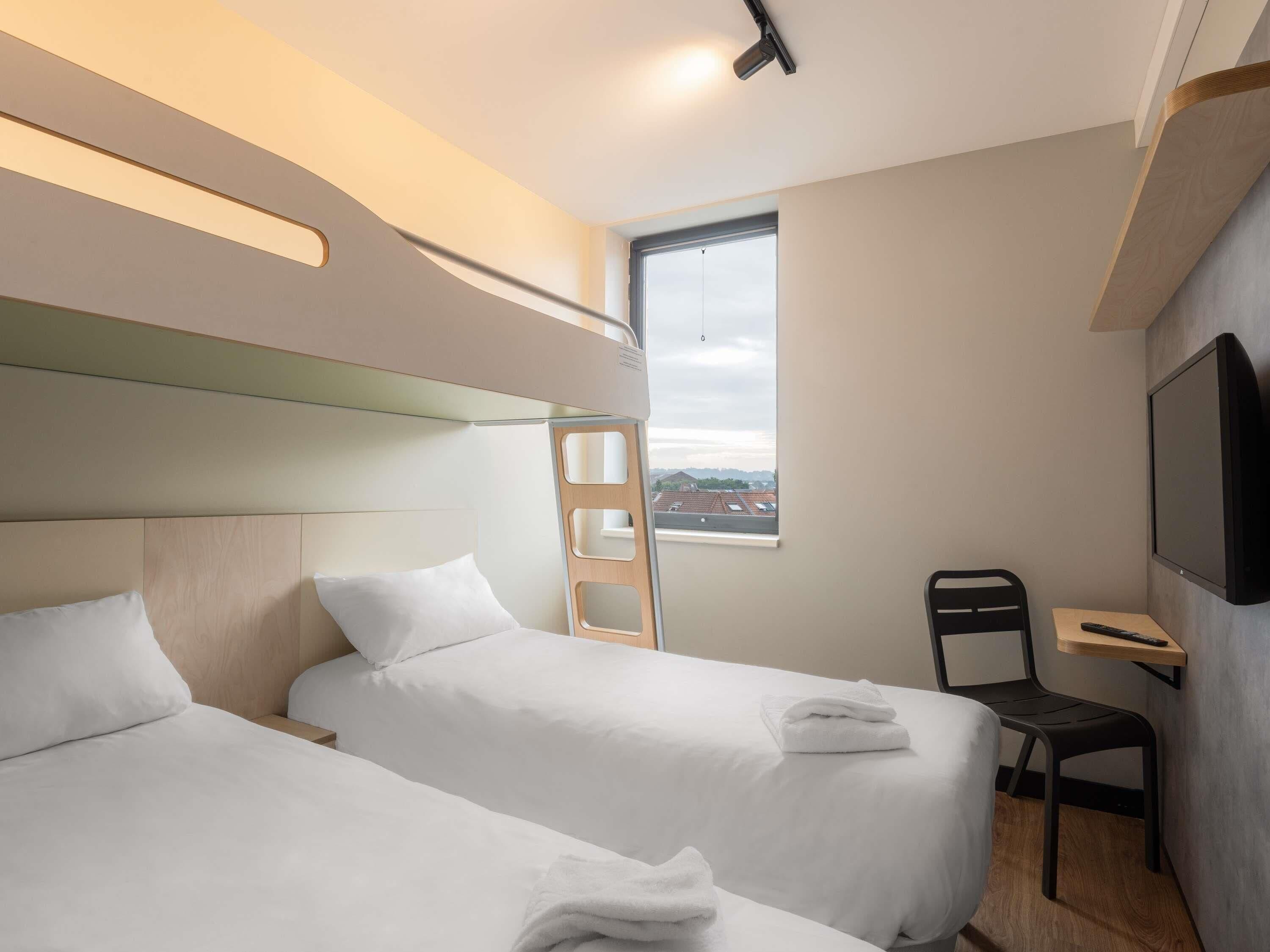 A modern hotel room with two beds, a TV, a desk, and a window with a city view.