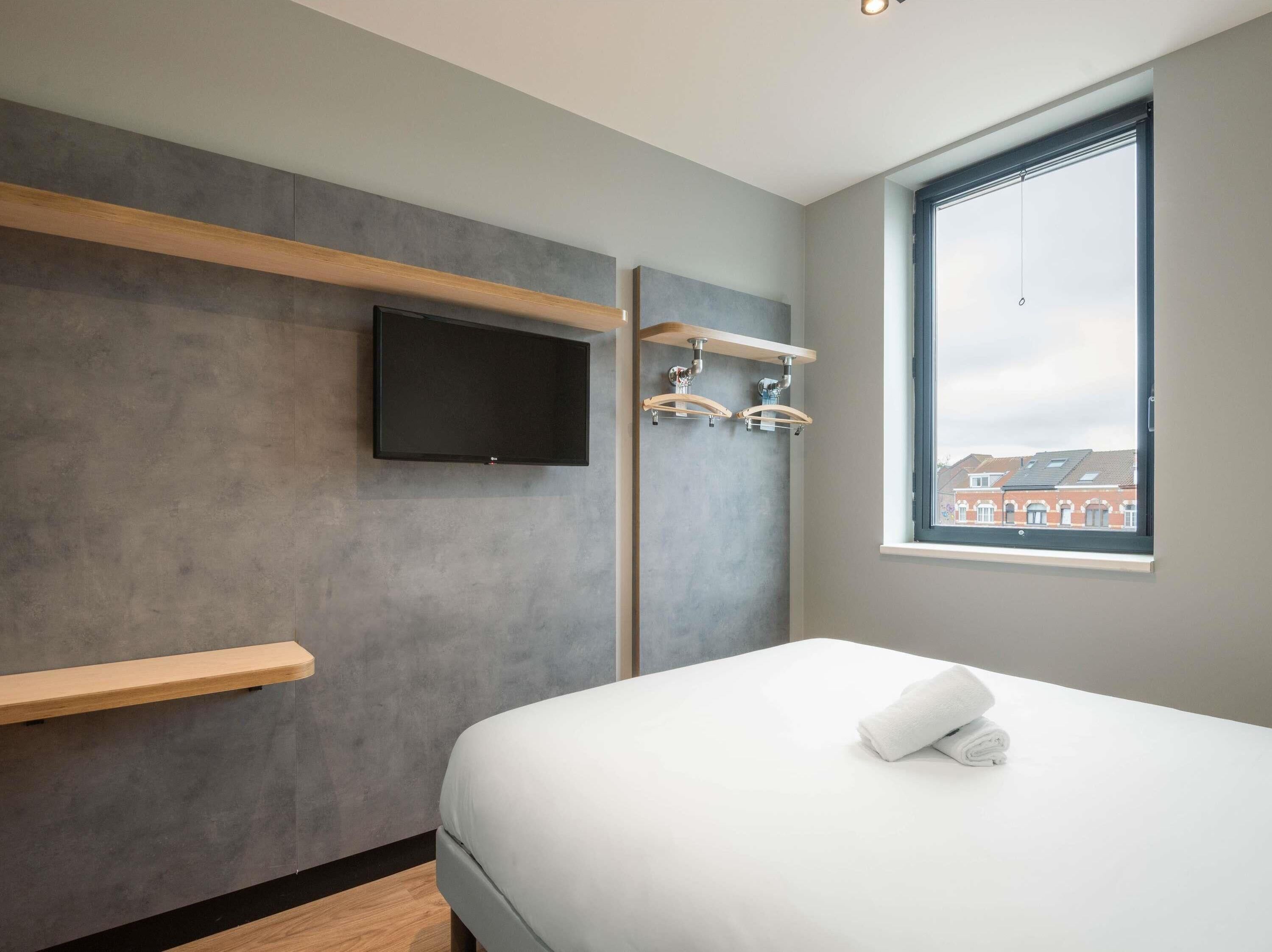 A modern hotel room with a bed, a mounted TV, and a window with a city view.