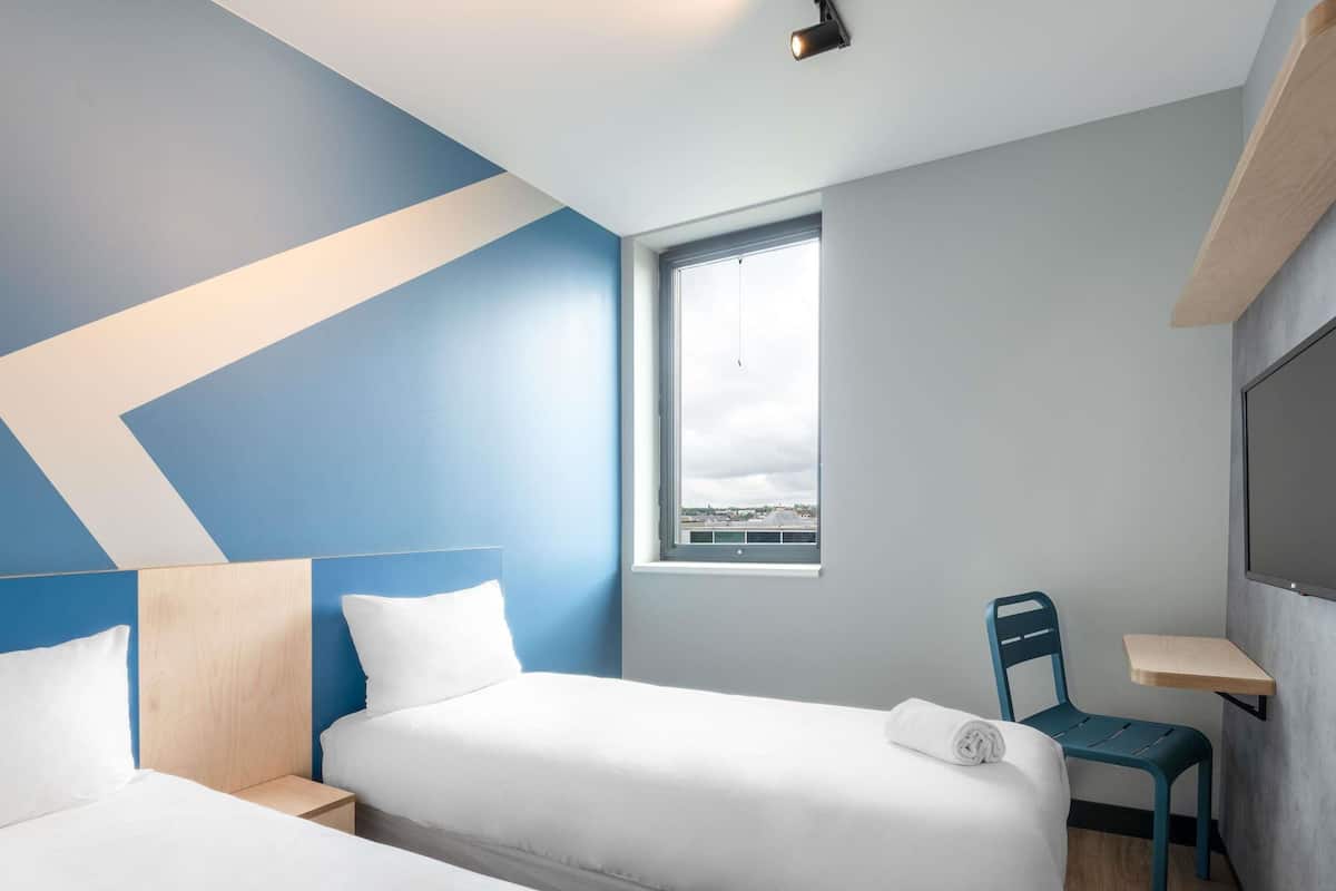 A modern hotel room with two beds, a large window, and a view of the cityscape.