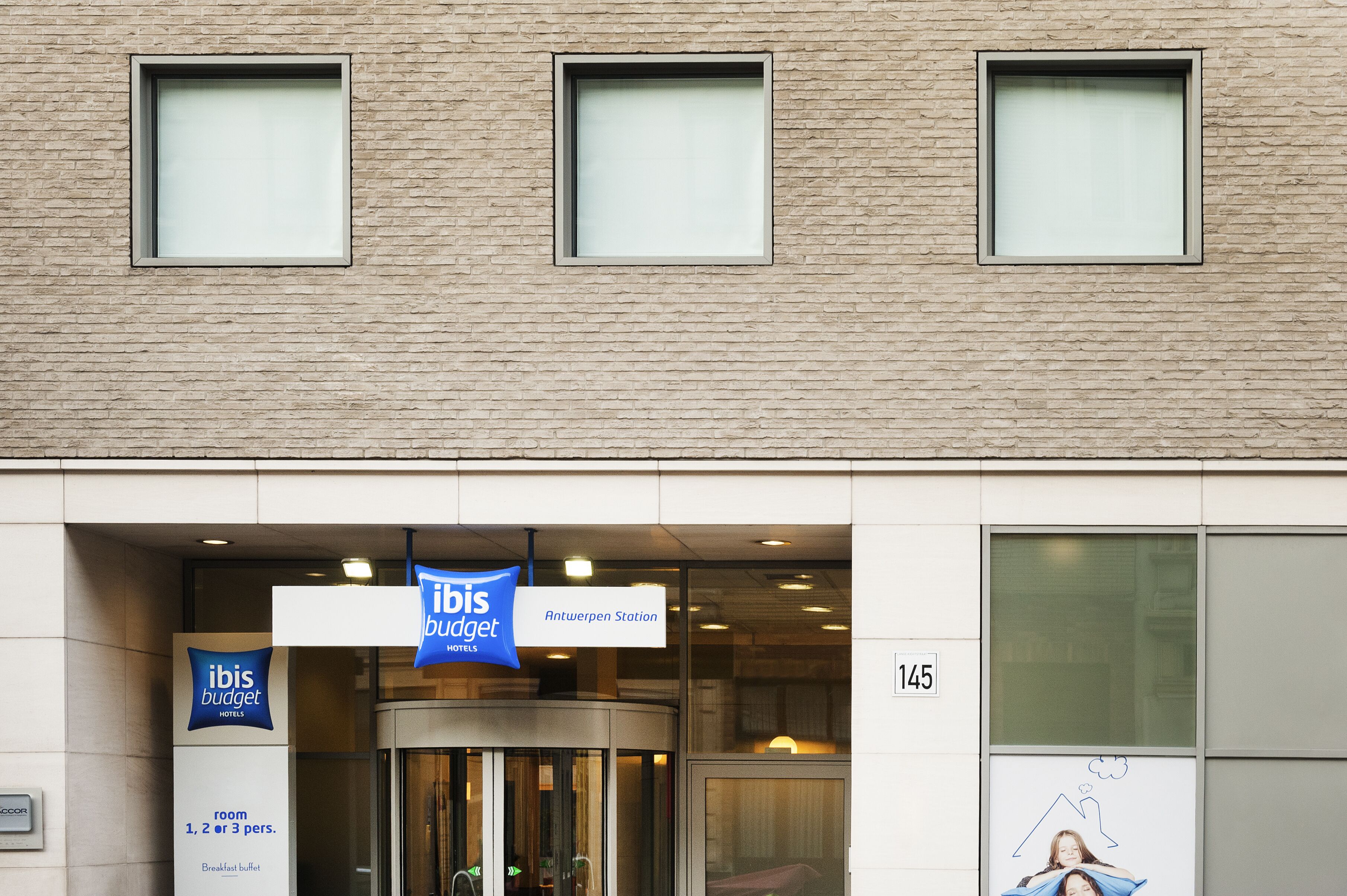 Entrada de un hotel con un cartel de ibis budget, una puerta giratoria y el número 145 en la pared.