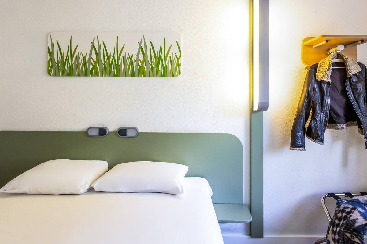 Une chambre d’hôtel comprenant un lit, une lampe fixée au mur, un porte-manteau avec des vestes et une œuvre d’art décorative accrochée au mur.