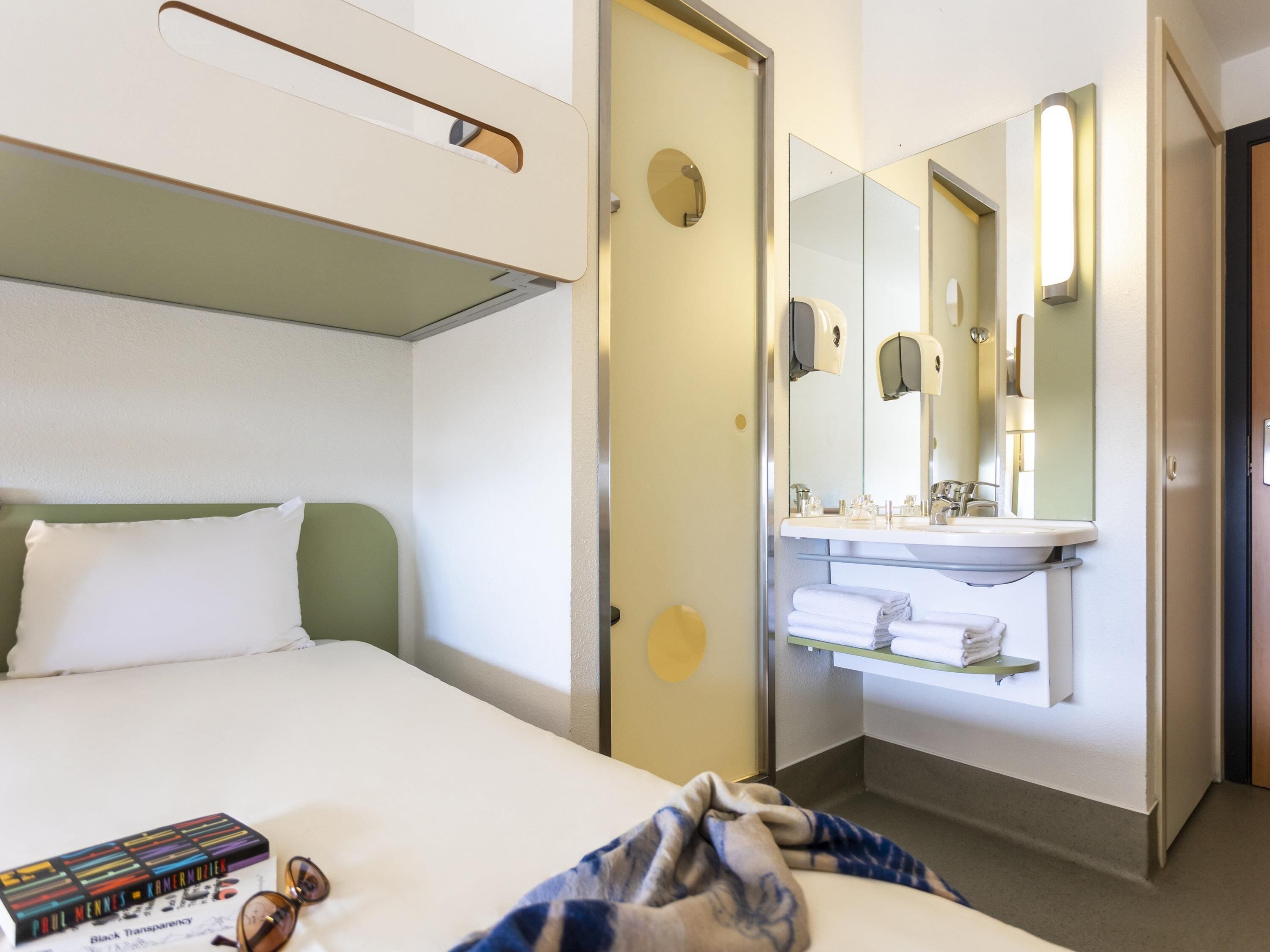 Una habitación de hotel con una cama, una mesita de noche con un libro y vasos, un baño con un lavabo y espejo, y una puerta que da a otra habitación.