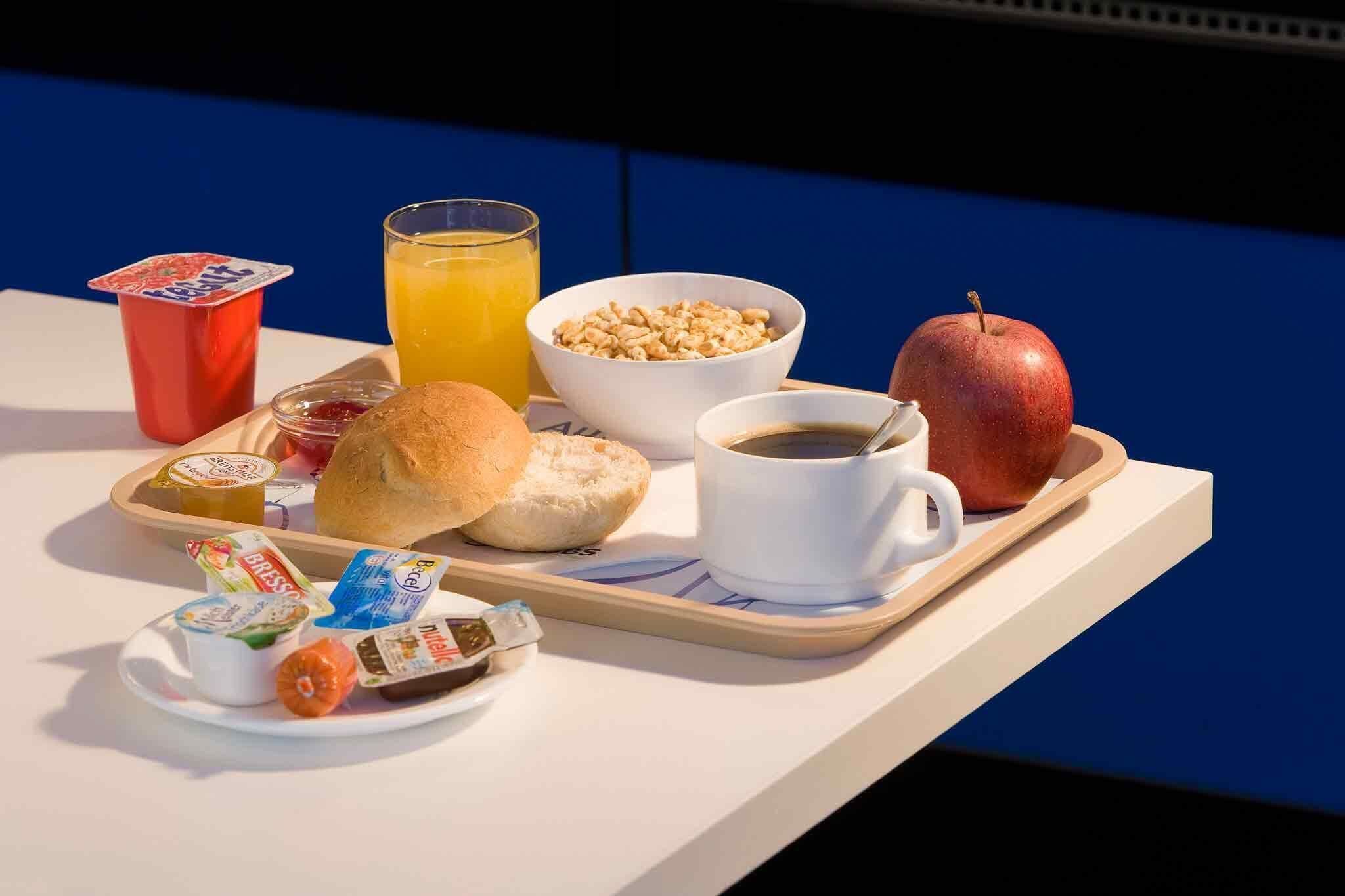 A breakfast tray with a croissant, apple, coffee cup, orange juice, yogurt, and cereal.