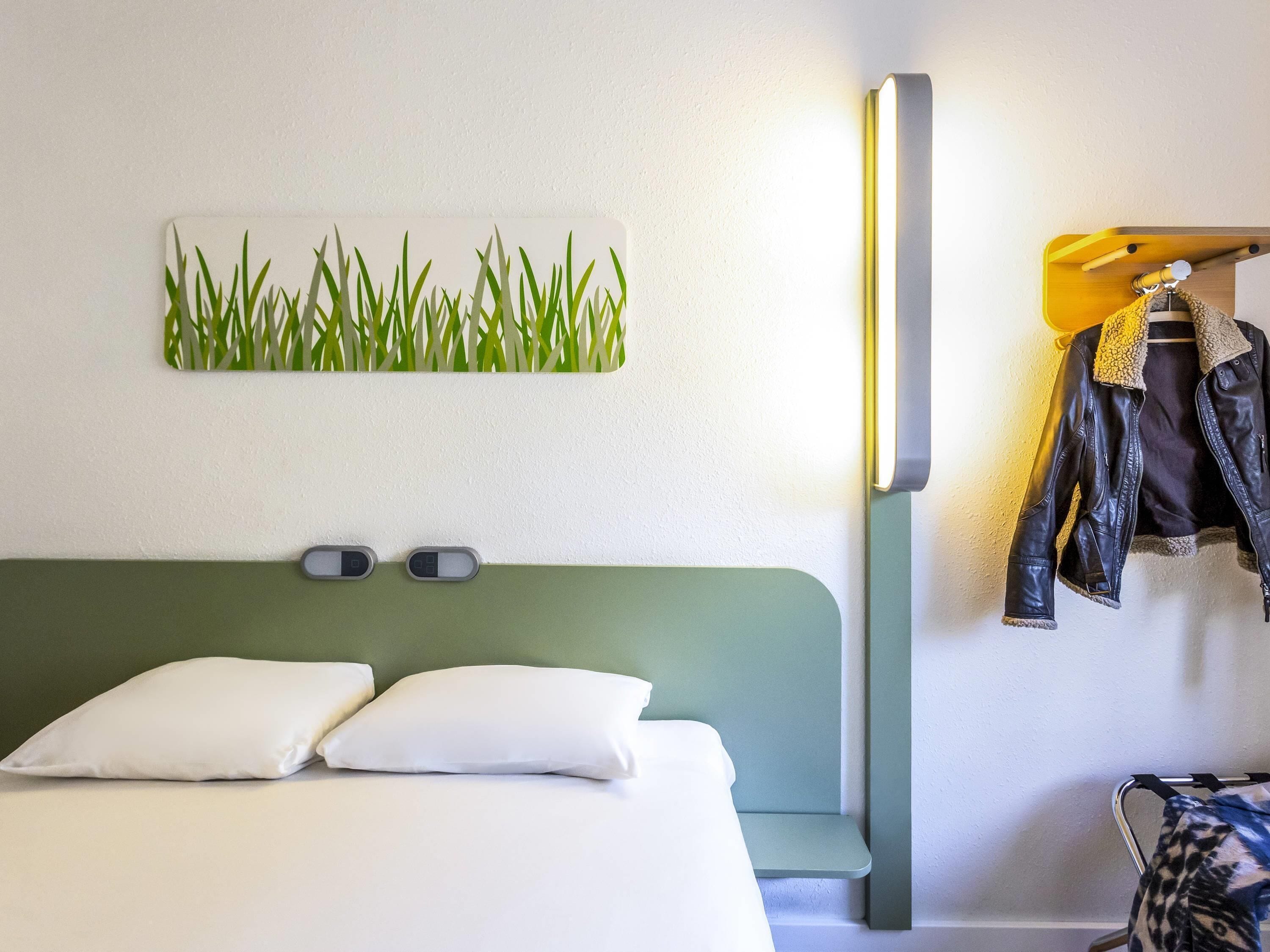 A hotel room with a bed, a wall-mounted light, and a coat rack with jackets.