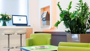 A reception area with a reception desk, a computer monitor displaying 'STAY CONNECT', a phone, and a plant. There are two green chairs and a table with magazines.