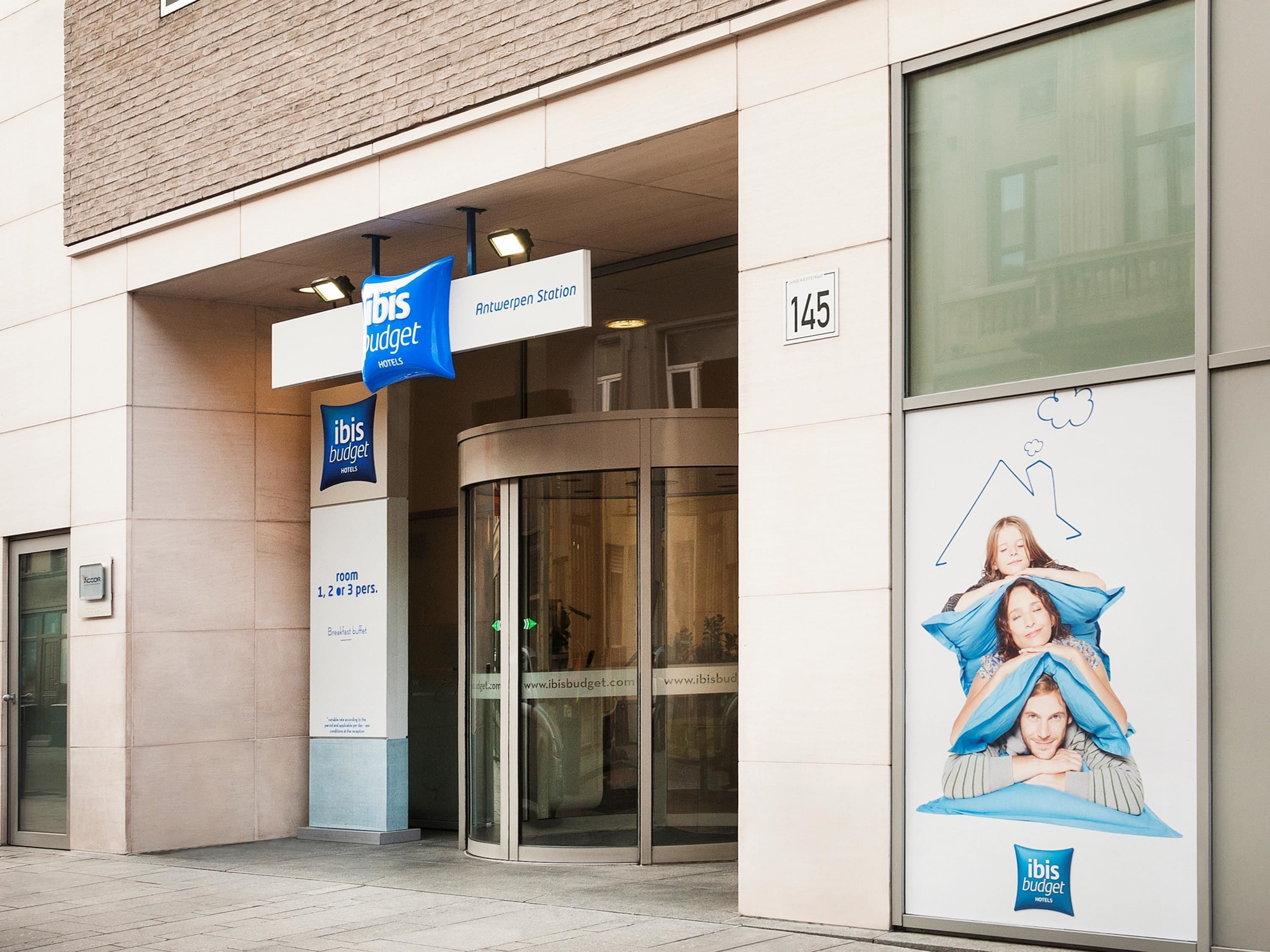 Entrada de un hotel con puerta de cristal, un letrero que indica 'Antwerpen Station' y un cartel grande que anuncia 'ibis budget hotels'.