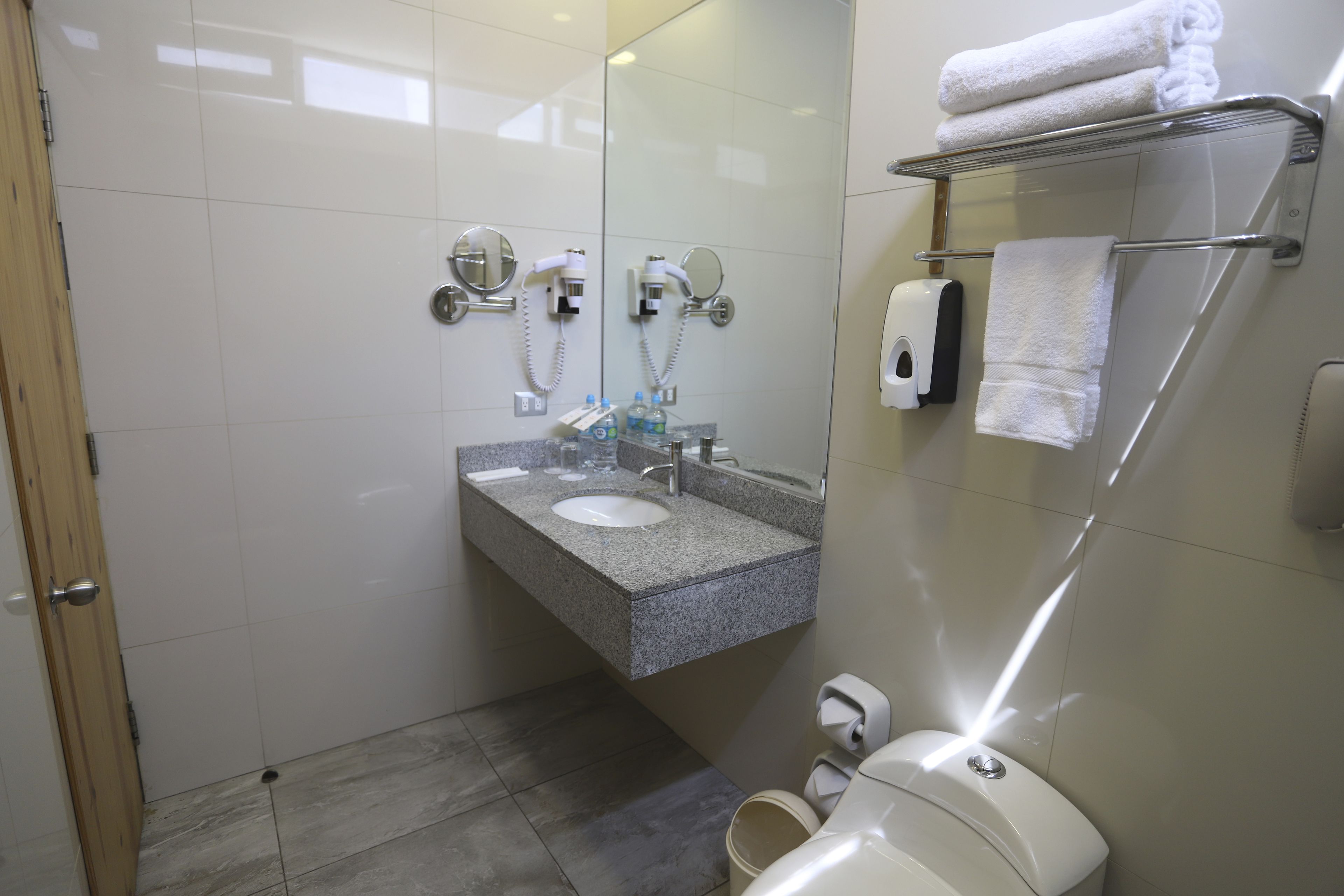 A bathroom with a granite countertop sink, a mirror, a towel rack with folded towels, a soap dispenser, and a toilet.