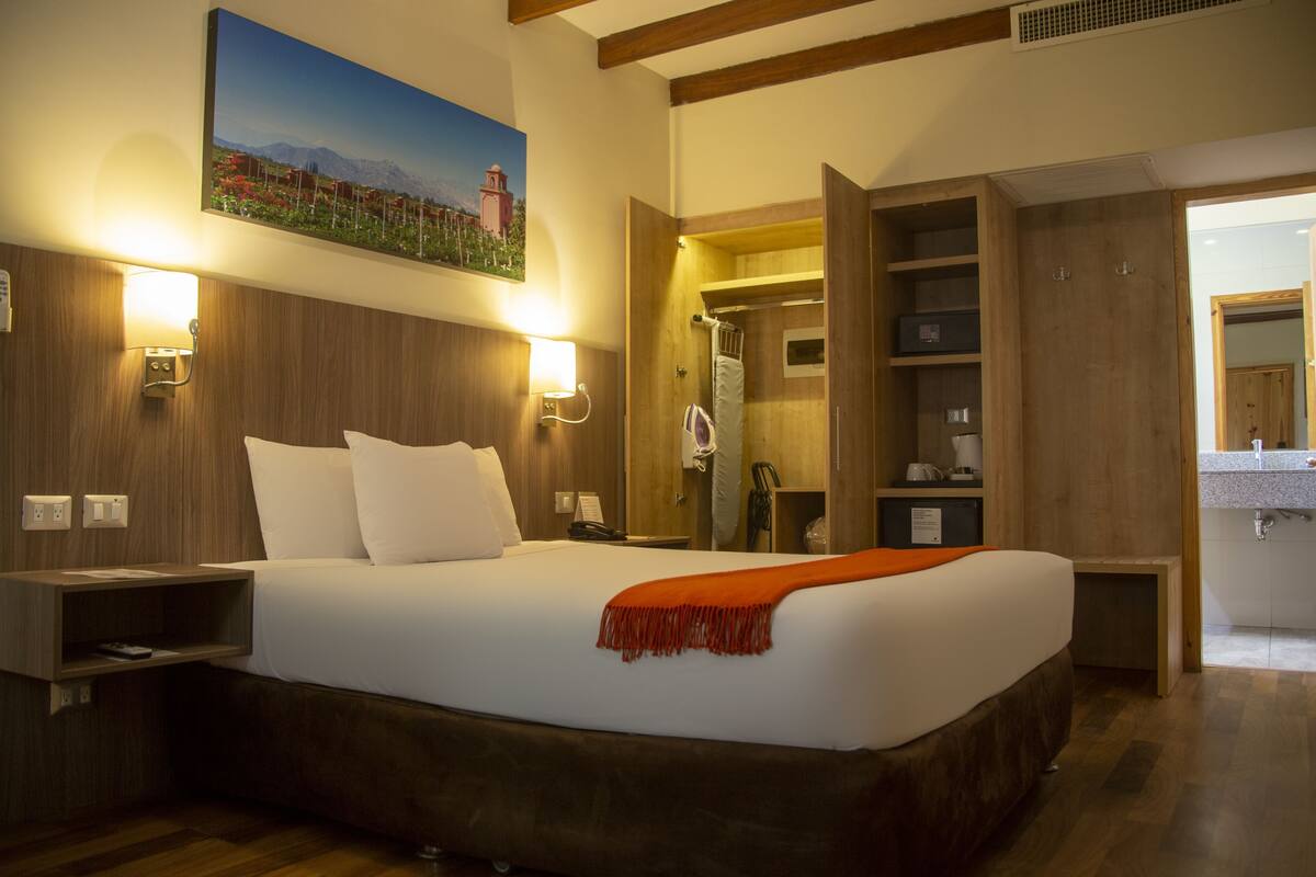 Una habitación de hotel con una cama, lámparas de noche, un cuadro, cabecera de madera y un armario de madera. Se ve un baño a través de una puerta abierta.