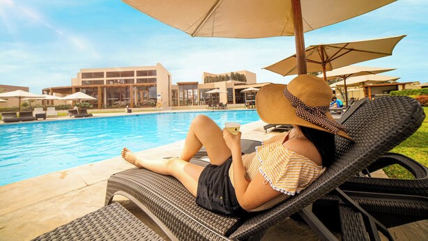 A poolside lounge area with wicker chairs and umbrellas, a pool, and modern buildings in the background. - Casa Andina Standard Chincha (Sunampe)