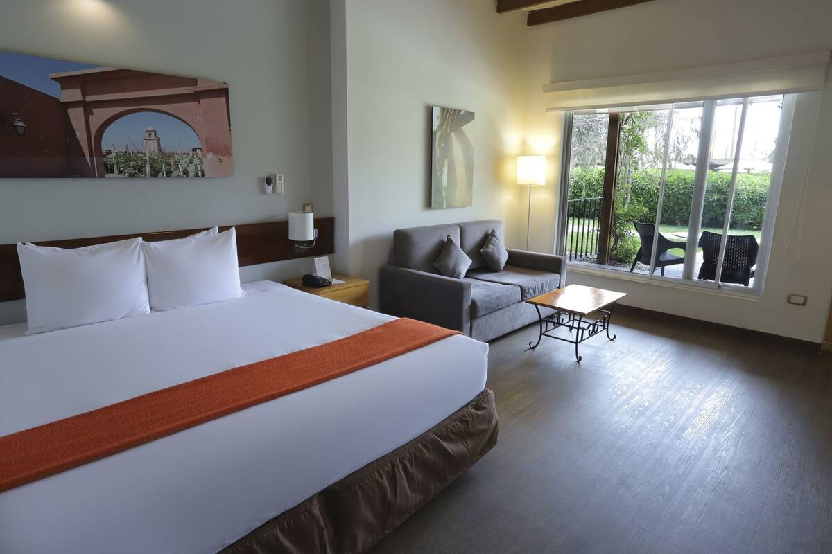 Habitación de hotel con una cama grande, un sofá, una mesita y un balcón con mobiliario exterior.