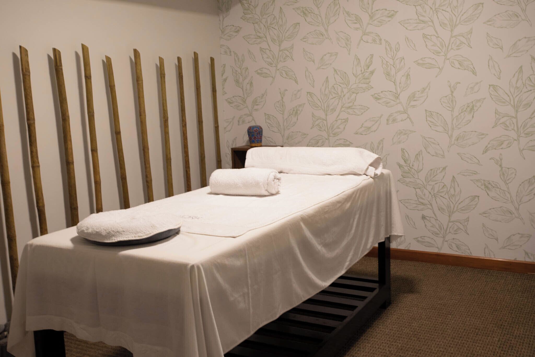 A single bed with a white sheet and a pillow, a small wooden stool with a vase, and a wall with a leafy pattern.