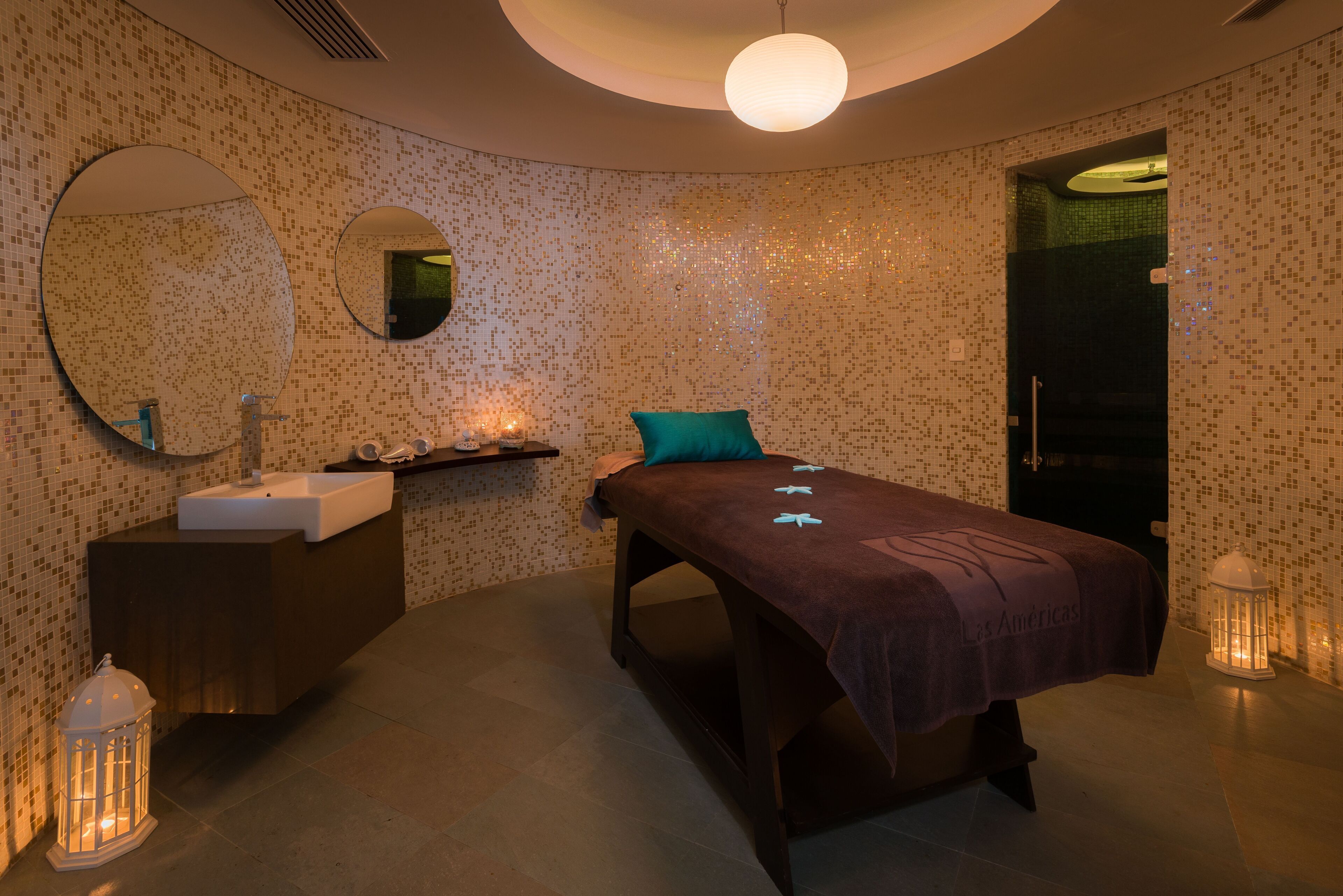 A modern spa room with a large mirror, a massage table, a sink, and a small shelf with candles.