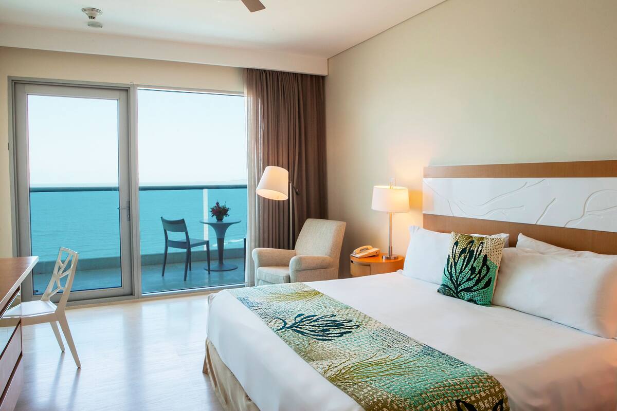 Una camera d'hotel con un letto, una scrivania, una sedia, un tavolo, una lampada e vista sull'oceano attraverso ampie finestre.